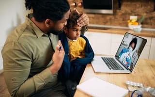 A parent sits with a child during a telemedicine appointment via video on a laptop.
